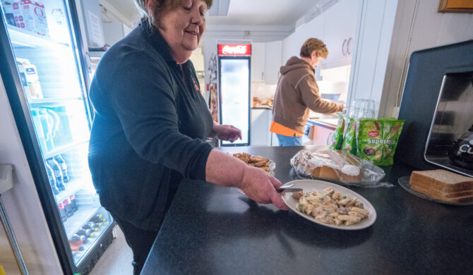 Bildet viser en dame som serverer en pastarett på en cafédisk. I bakgrunnen holder en annen dame på med å ordne noe på kjøkkenet.