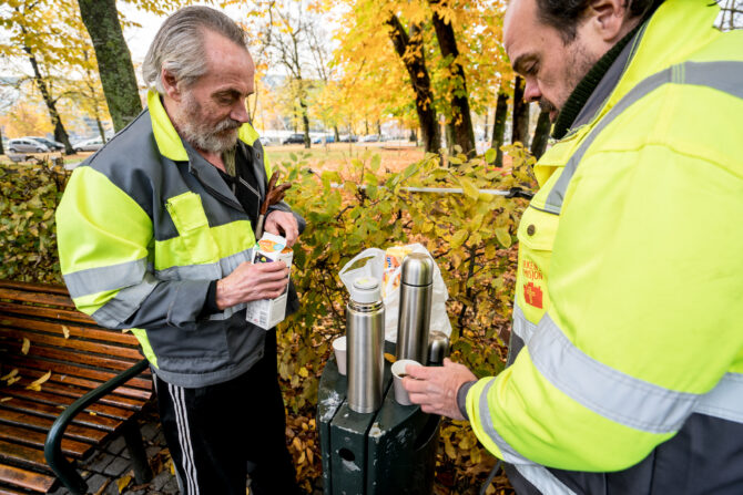 Bildet viser to menn i gule refleksvester som står i en park med trær i gule høstfarger bak seg og er i ferd med å helle kaffe fra termos i kopper.