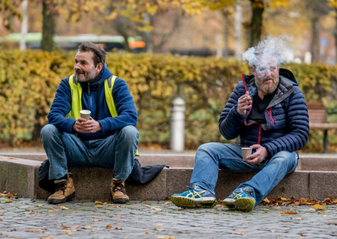 Bildet viser to menn som sitter på kanten av en tørrlagt fontene på høsten. Mannen til venstre har blå genser og gul refleksvest på seg. Han ser ut mot venstre i bildet. Mannen til høyre har blå boblejakke og sitter og tar seg en røyk mens han ser ut mot høyre av bildet.