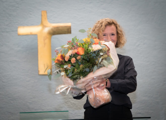 Bildet viser Kari Veiteberg med en stor blomsterbukett foran seg. Ansiktet hennes er halvveis skjult av blomstene. På veggen bak henne henger det et stort kors i gull