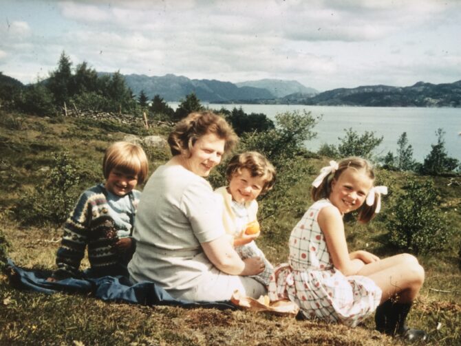 Bildet viser et eldre fargefotografi av en familie som sitter ute i naturen på grønt gress. I bakgrunnen skimtes havet og blå fjelltopper enda lenger bak. Det er moren til Kari Veiteberg som sitter forrerst i bildet, og Veiteberg og to av hennes søstre som barn som sitter sammen med henne.