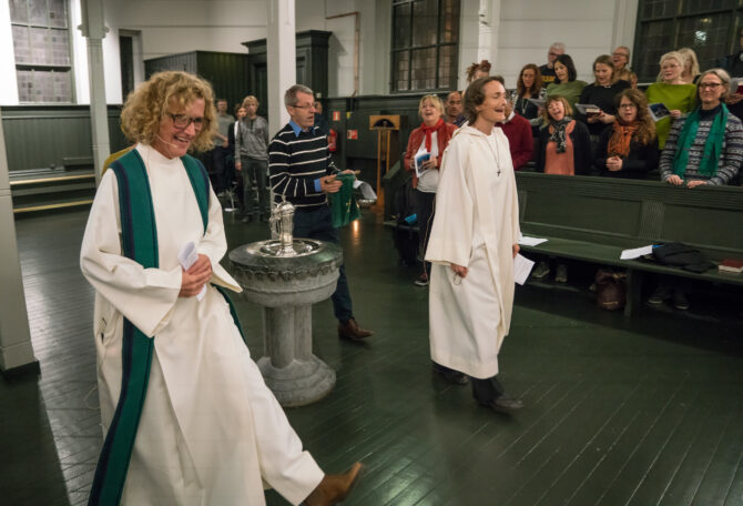 Bildet er fra kirkerommet i andre etasje i Tøyenkirken i Oslo. Kari Veiteberg (foran i bildet) går i hvit prestekjole sammen med to andre gjennom kirkerommet. I bakgrunnen sees kirkegjengere i benkeradene som står og synger med.