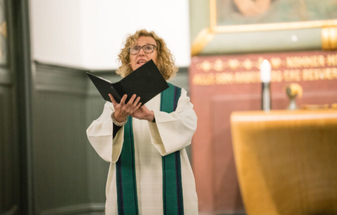 Bildet viser Kari Veiteberg i hvit prestekjole i et kirkerom. Hun holder en svart bok med tekst hun leser fra på hendene sine, med hendene høyt hevet.