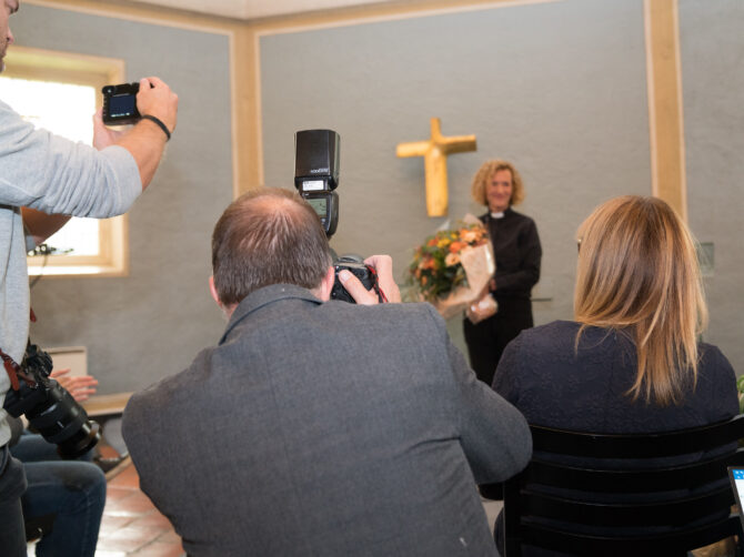Bildet viser en gjeng med fotografer i forgrunnen med kameraene rettet mot Kari Veiteberg, Oslos nye biskop, som står i bakgrunnen av bildet. På veggen bak henne skimtes et kors av gull.