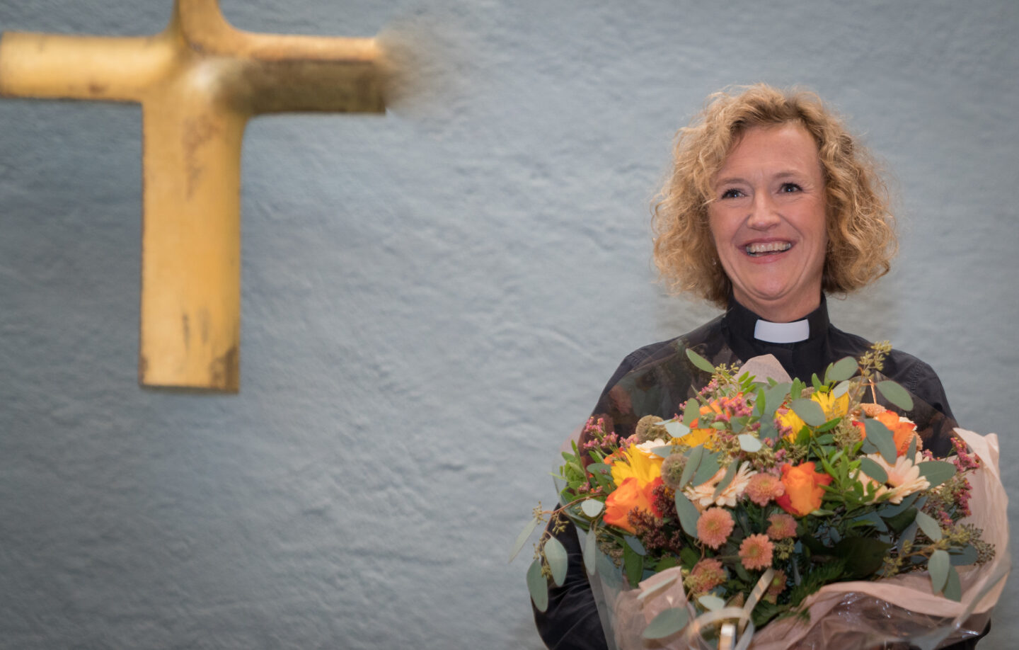 Bildet viser Kari Veiteberg stående med en stor blomsterbukett i armene. Hun smiler bredt og har på seg en svart skjorte med prestekrage. Bak henne på veggen til venstre henger et stort kors i gull.