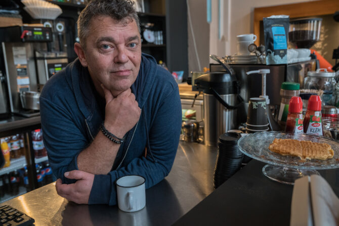 Bildet viser Trond Henriksen som lener seg over en cafédisk mens han ser mot fotografen. Han lener haken under hånda si, og har en kaffekopp foran seg.