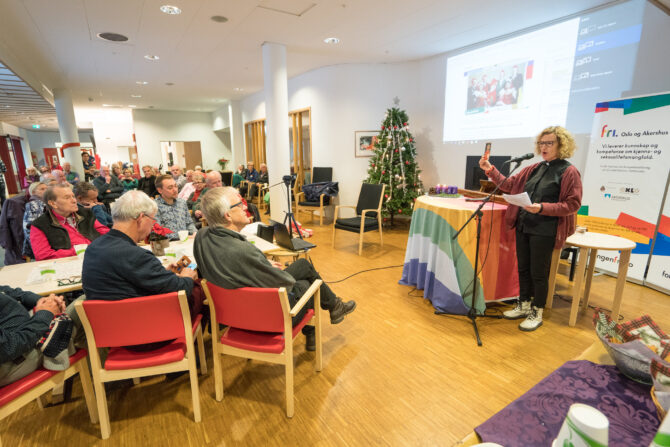 Bildet viser et forsamlingslokale med eldre mennesker på røde stoler og en kvinne som står til høyre ved et podie som er dekket av et regnbueflagg. Kvinnen heter Kari Veiteberg og er avbildet tre dager før hun blir vigslet som Oslos nye biskop.