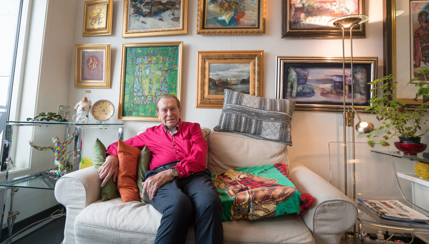 Bildet viser en eldre mann i rosa skjorte som sitter i en hvit sofa i en leilighet på Kampen Omsorg+. På veggene rundt ham henger det masse kunstverk i flotte rammer.