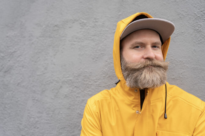 Adam i gul regnjakke som smiler lurt foran en grå sementvegg.